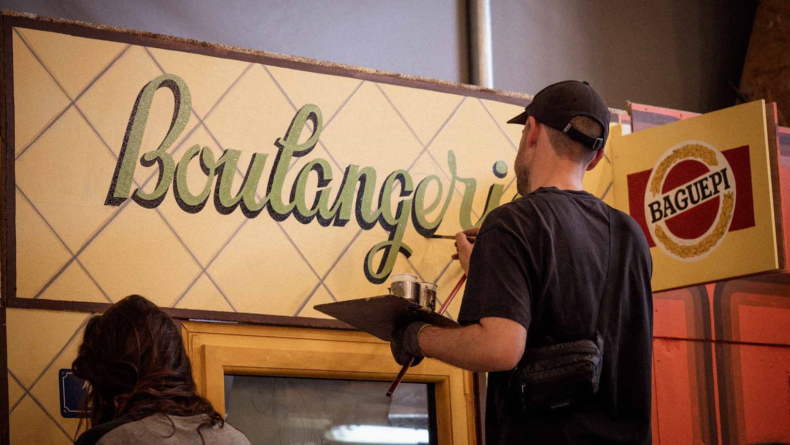 View from behind a man painting a shopfront that says "Boulangerie"