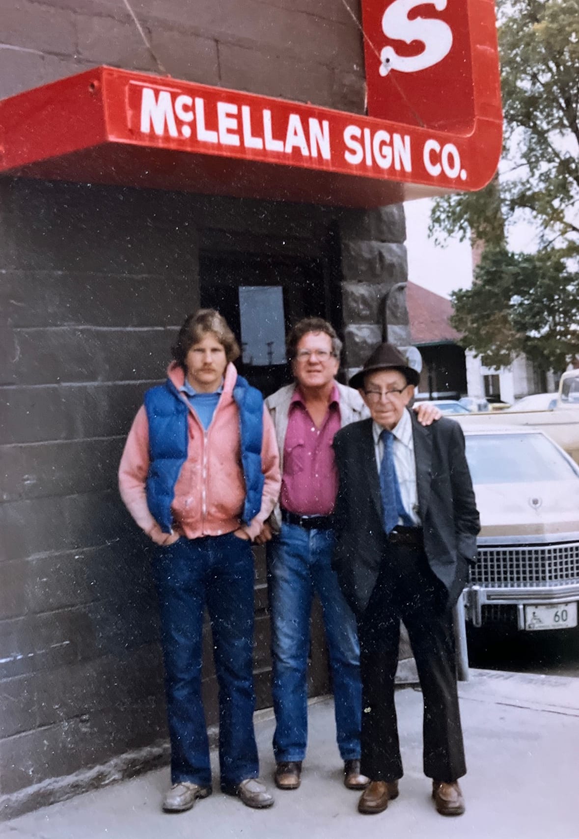 Three men posing for a portrait in front of a corner shop with white lettering on a red sign that read "McLellan Sign Co.".