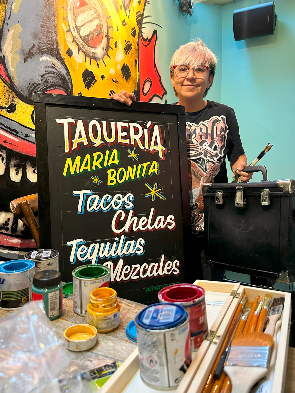 Woman posing in an artists' studio setting with a hand-painted blackboard for a taquer¡a.