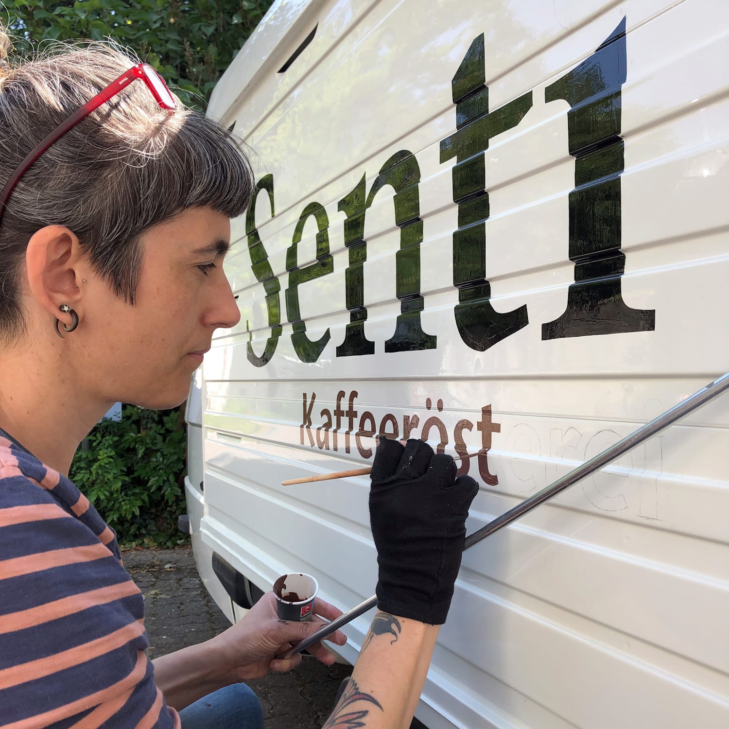 Woman resting her gloved hand on a mahl stick as she paints lettering on the corrugated side of a vehicle.