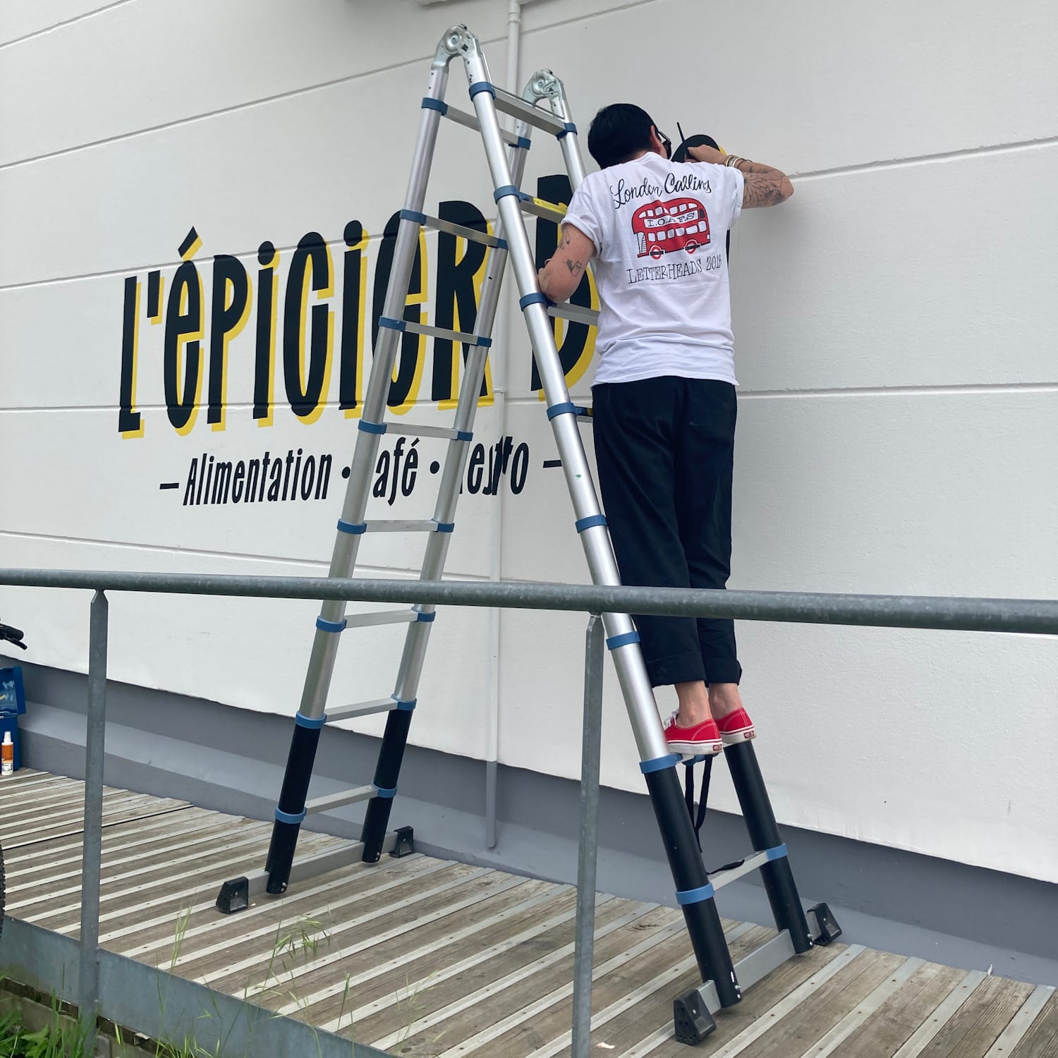 Woman on the second rung of a ladder painting black lettering with a yellow shade onto a wall.