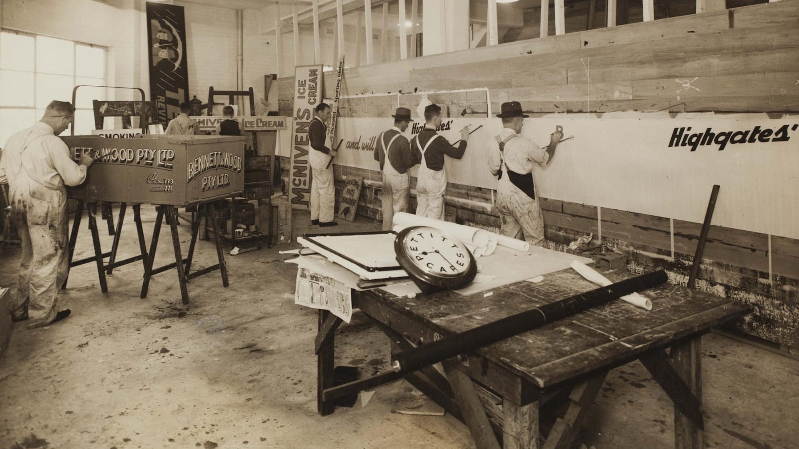 Black and white photo of a sign painting workshop setting with men at work on a variety of projects on an easel.