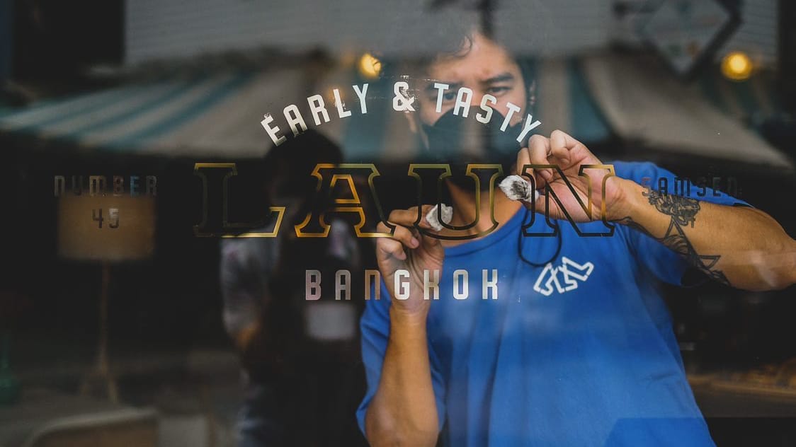 Man photographed through a shop window where he is working on a painted and gilded sign that reads "Early & Tasty, Laun, Bangkok".