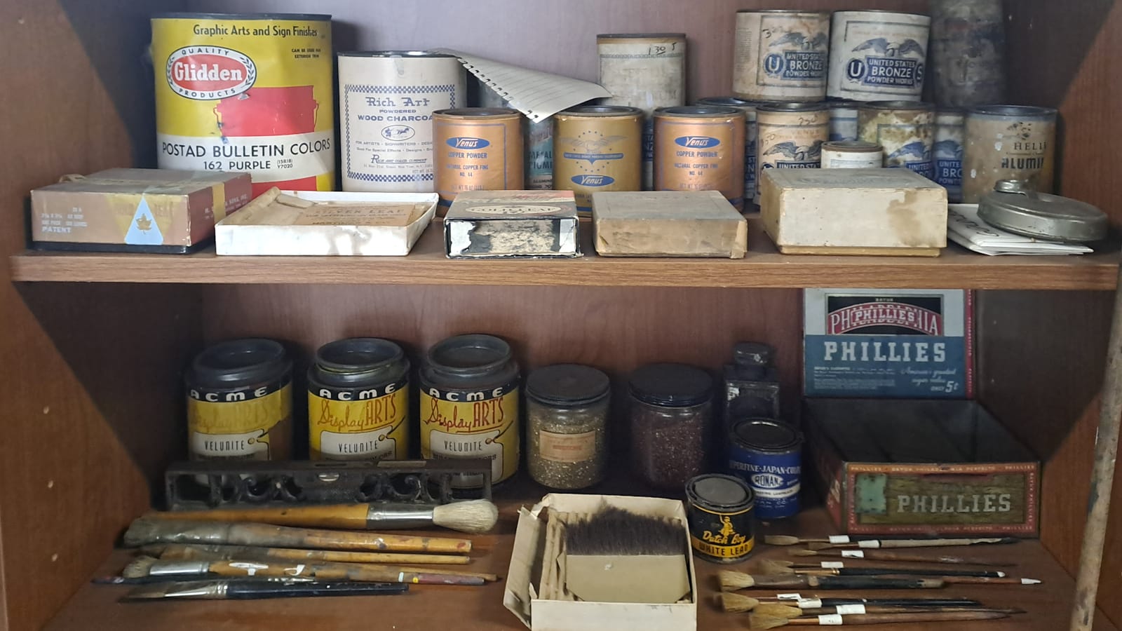 Two shelves filled with an assortment of paint tins, brushes, boxes, and other items.