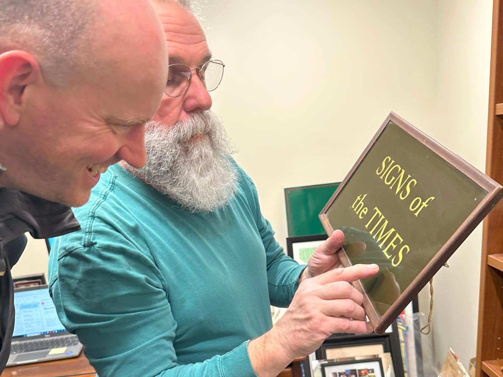 Two men looking at a gilded glass sign in a frame. It says "Signs of the Times".