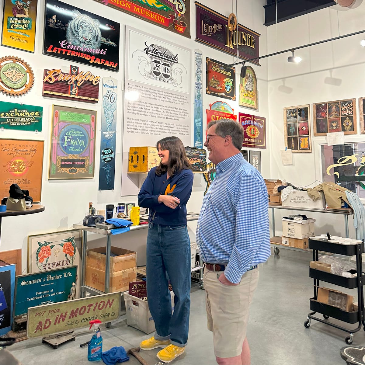 Two people smiling in a gallery/museum setting with hand-painted signs mounted from floor to ceiling.