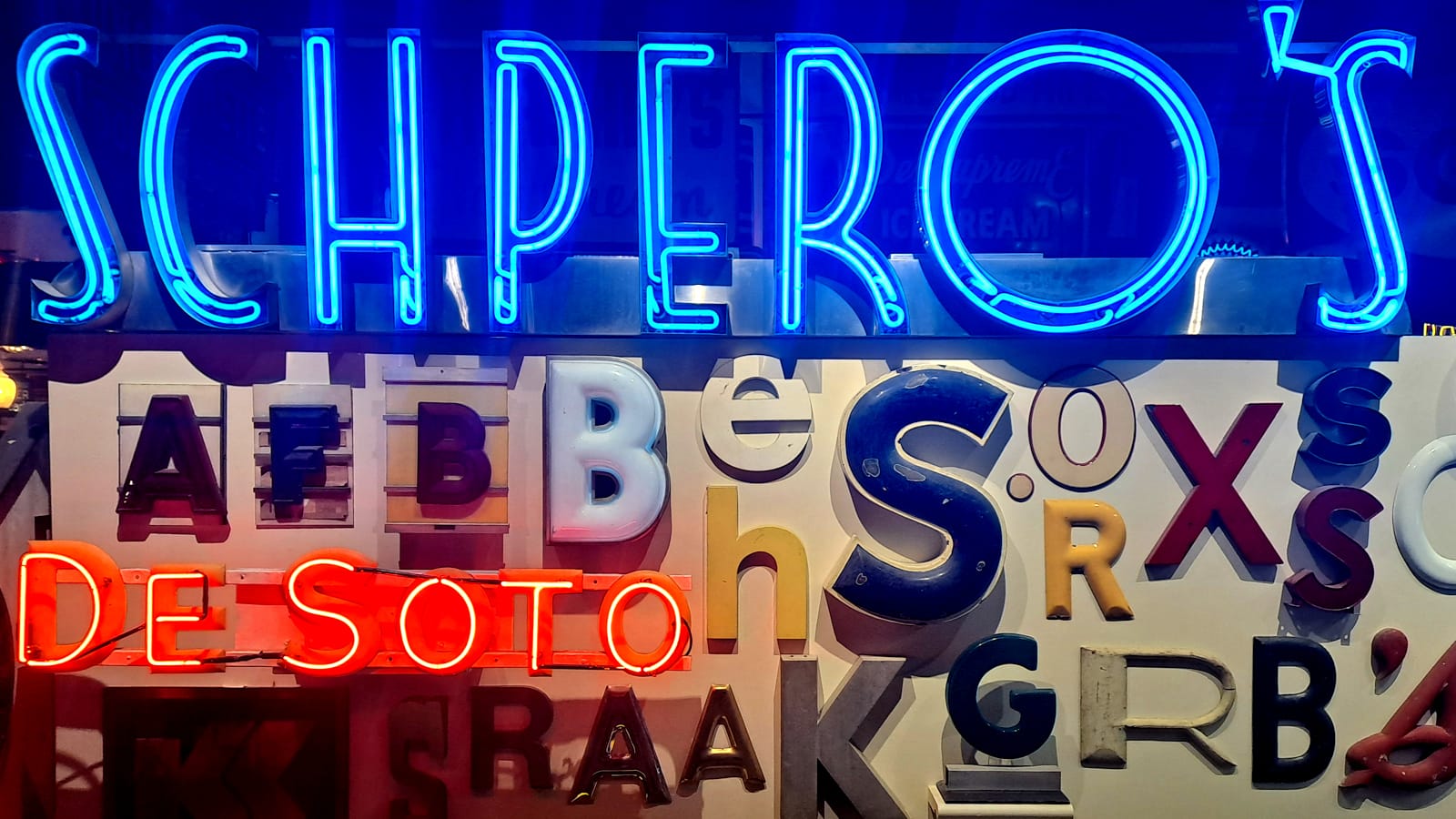 Array of letters mounted to a wall. Above this is an illuminated blue neon sign in Art Deco letters for "Schpero's" and amongst them another neon sign in red that says "De Soto".