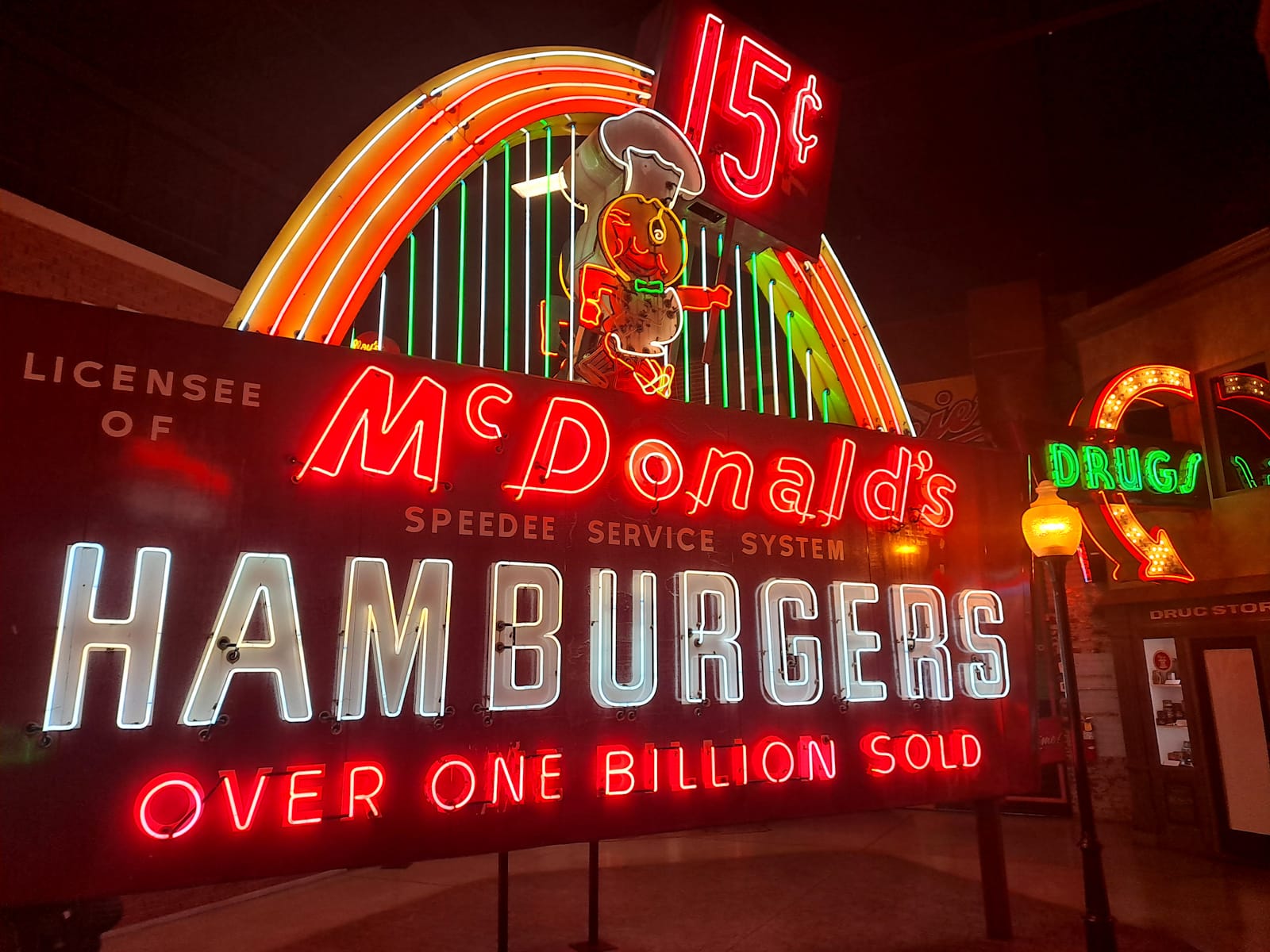 Huge neon sign for McDonald's that boasts of "Over One Billion Sold" and has a chef-hatted mascot at the top holding a sign that says "15c". Slight set back from this is another illuminated neon sign comprising a curved arrow and the single word "Drugs".