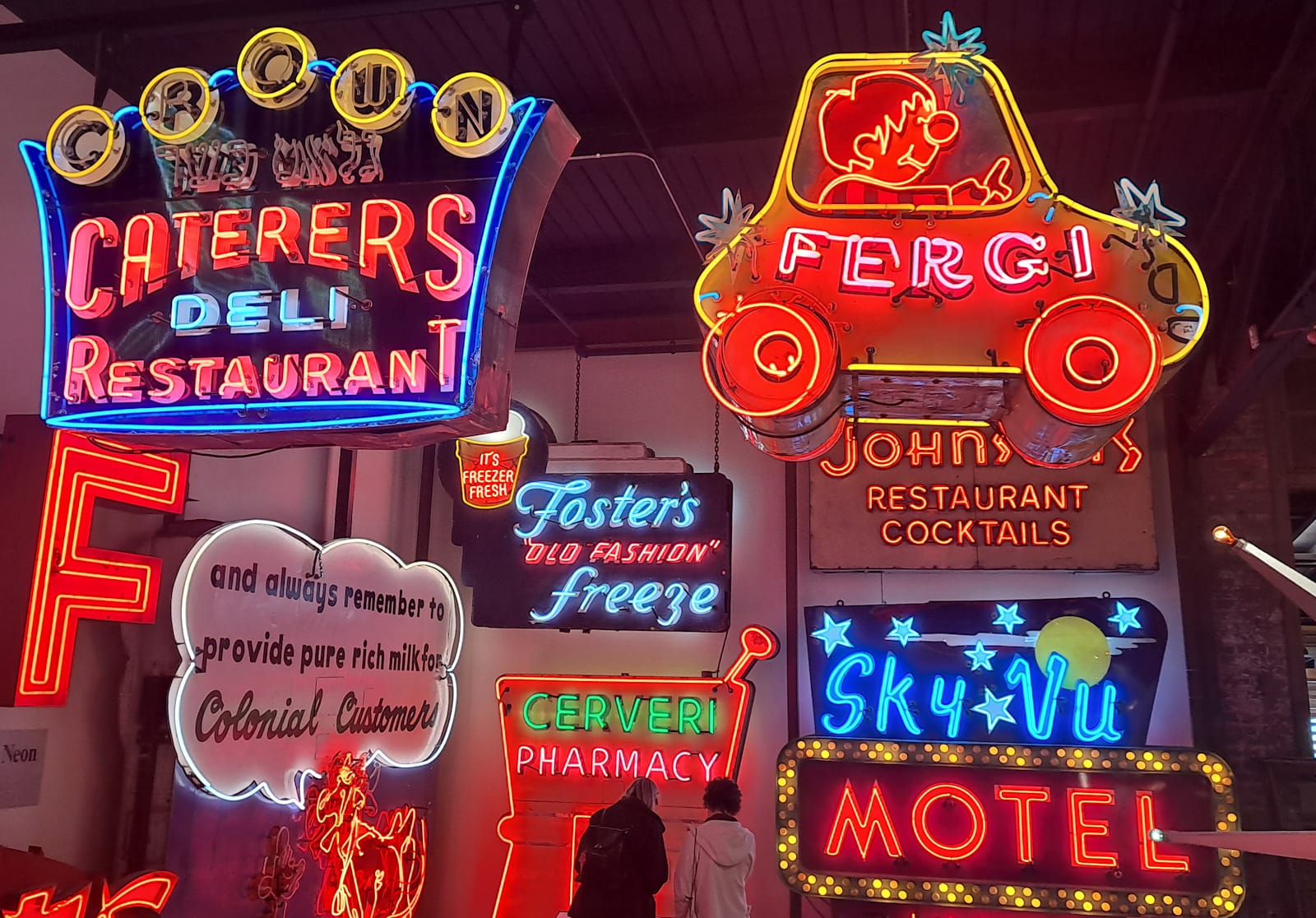 Medley of illuminated neon signs mounted in a gallery/museum setting, some to the walls and some from the ceiling.