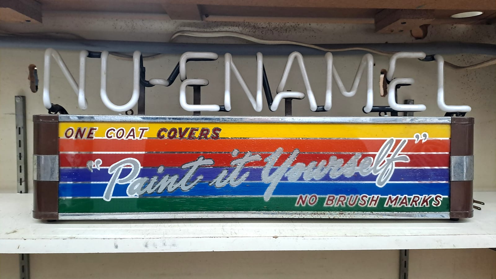 Small cabinet with unlit neon letters above that say "Nu-Enamel". Below this are letters on a horizontal rainbow background that say "One Coat Covers, "Paint it Yourself", No Brush Marks".