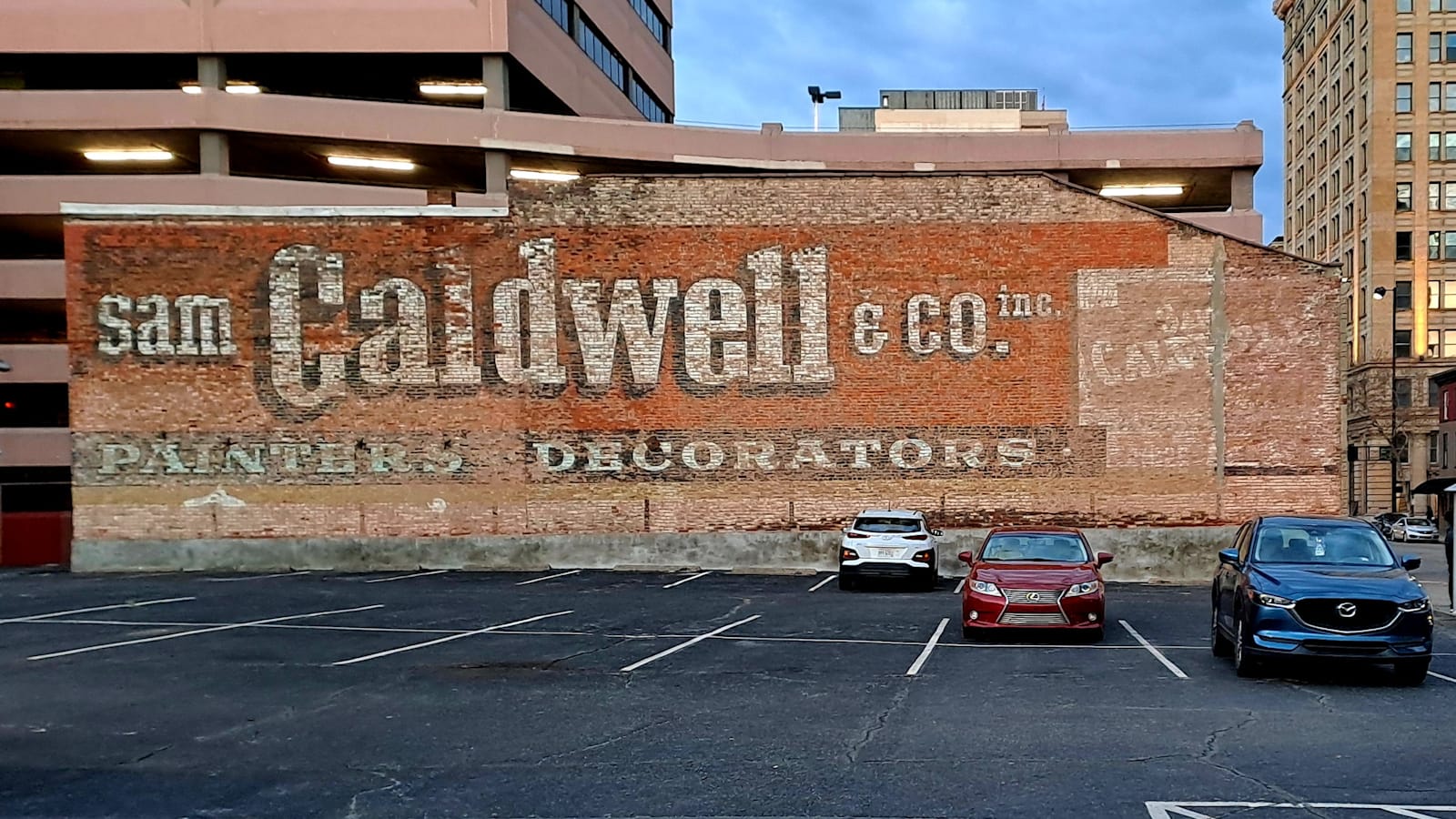 Large brick wall facing a parking lot with a fading hand-painted sign for Sam Caldwell & Co, Painters, Decorators. 