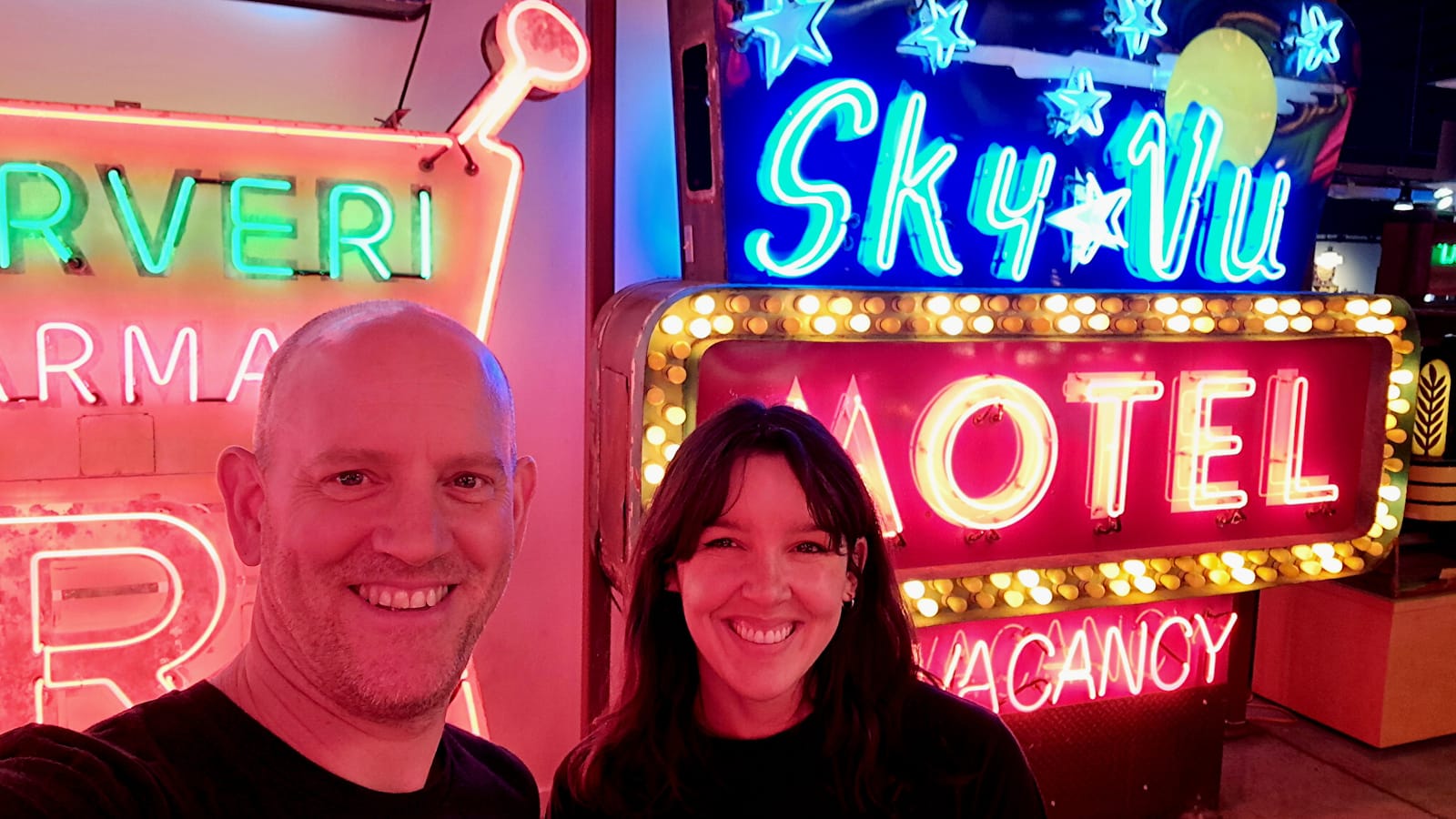 Two people smiling for a selfit in front of an illuminated neon sign for the Sky Vu Motel.