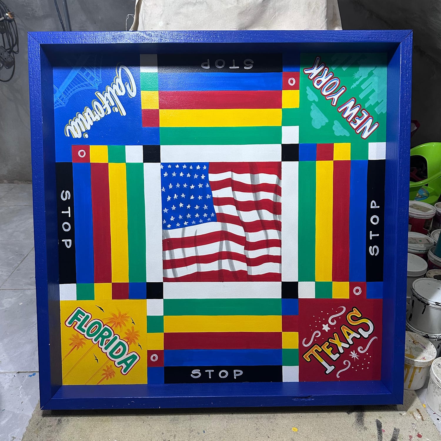 Stylised Ludo board with each of the home bases being lettered in styles representative of their locations: Florida; Texas; New York; and California. The USA flag is set in the central square with the squares for moving around the board painted in alternating squares and rectangles in white, green, yellow, red, blue, and black.