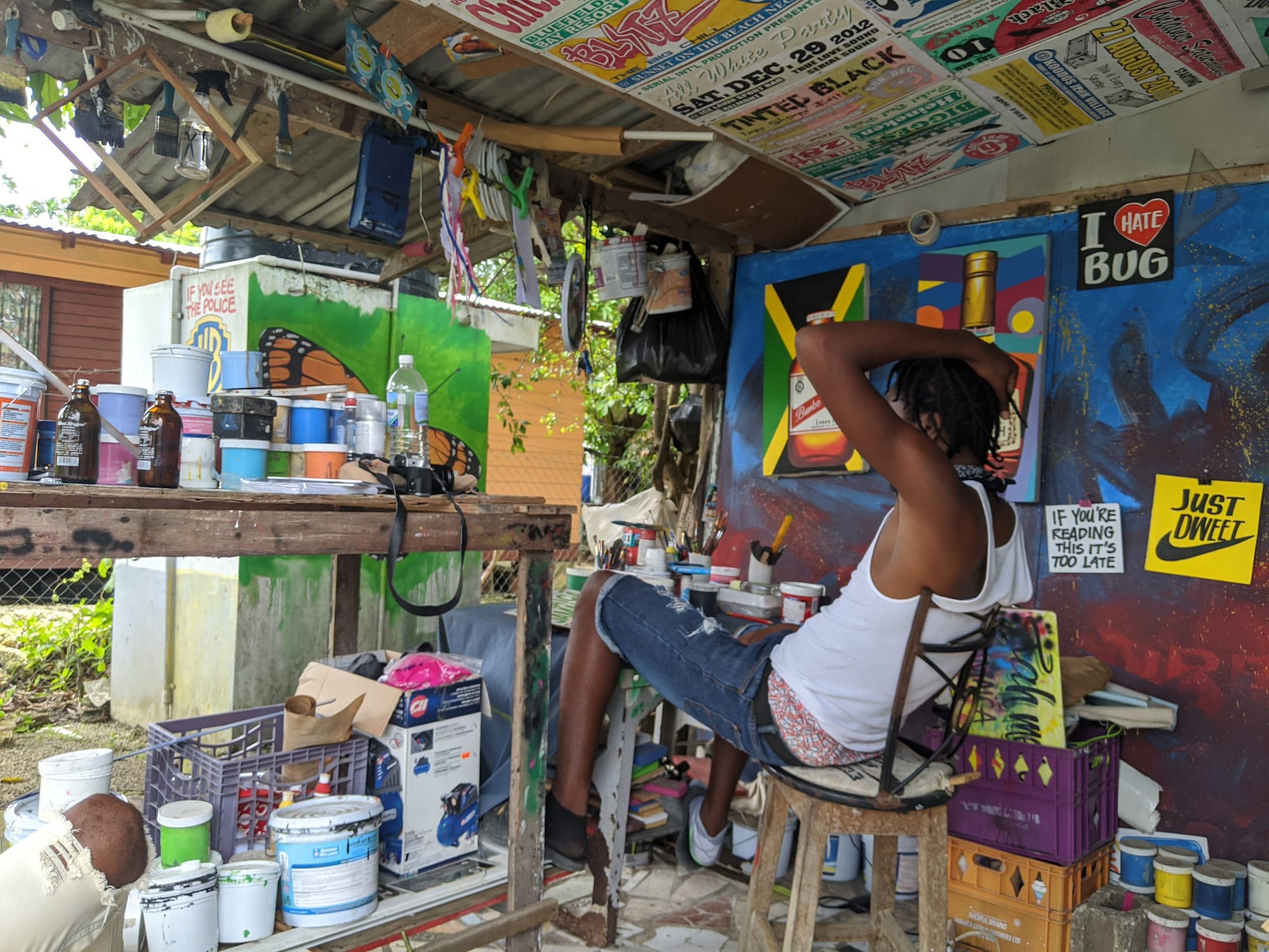 Main say on a stool with his hand resting on his head as he looks away from the camera out out of his open air painter's studio.