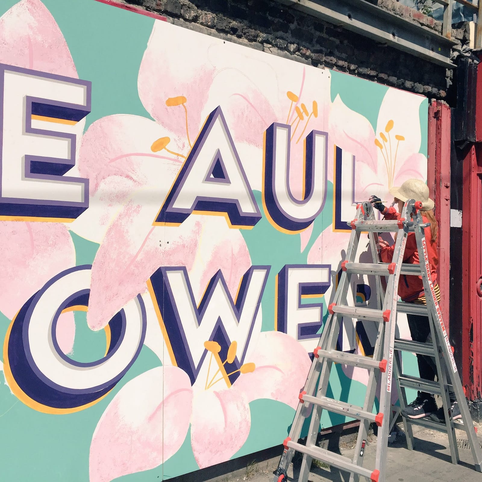 Sign painter in a hat painting onto a wall with some ladders between them and the viewer.