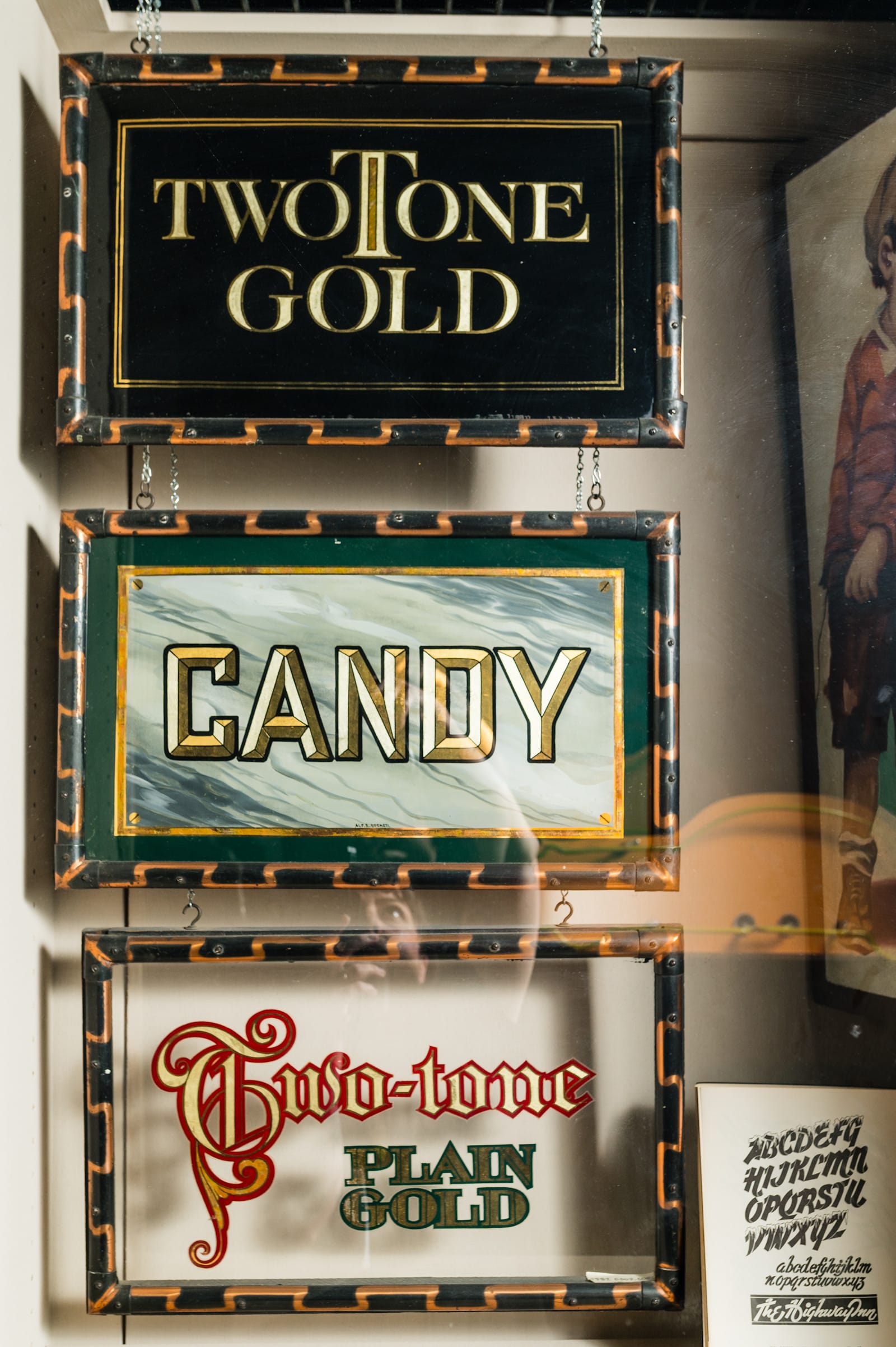 Gilded panels displayed vertically inside a museum cabinet. The top one says "TwoTone Gold" in high contrast serifed lettering in two tones of gold on a black background. Below this is "Candy" in prismatic lettering achieved by the use of different gilding effects and all set on a faux marbled background. The bottom panel says "Two-tone, Plain Gold" with two-tone in gilded blackletter with a red outline and plain gold in subtly slab-serifed letters with a green outline.