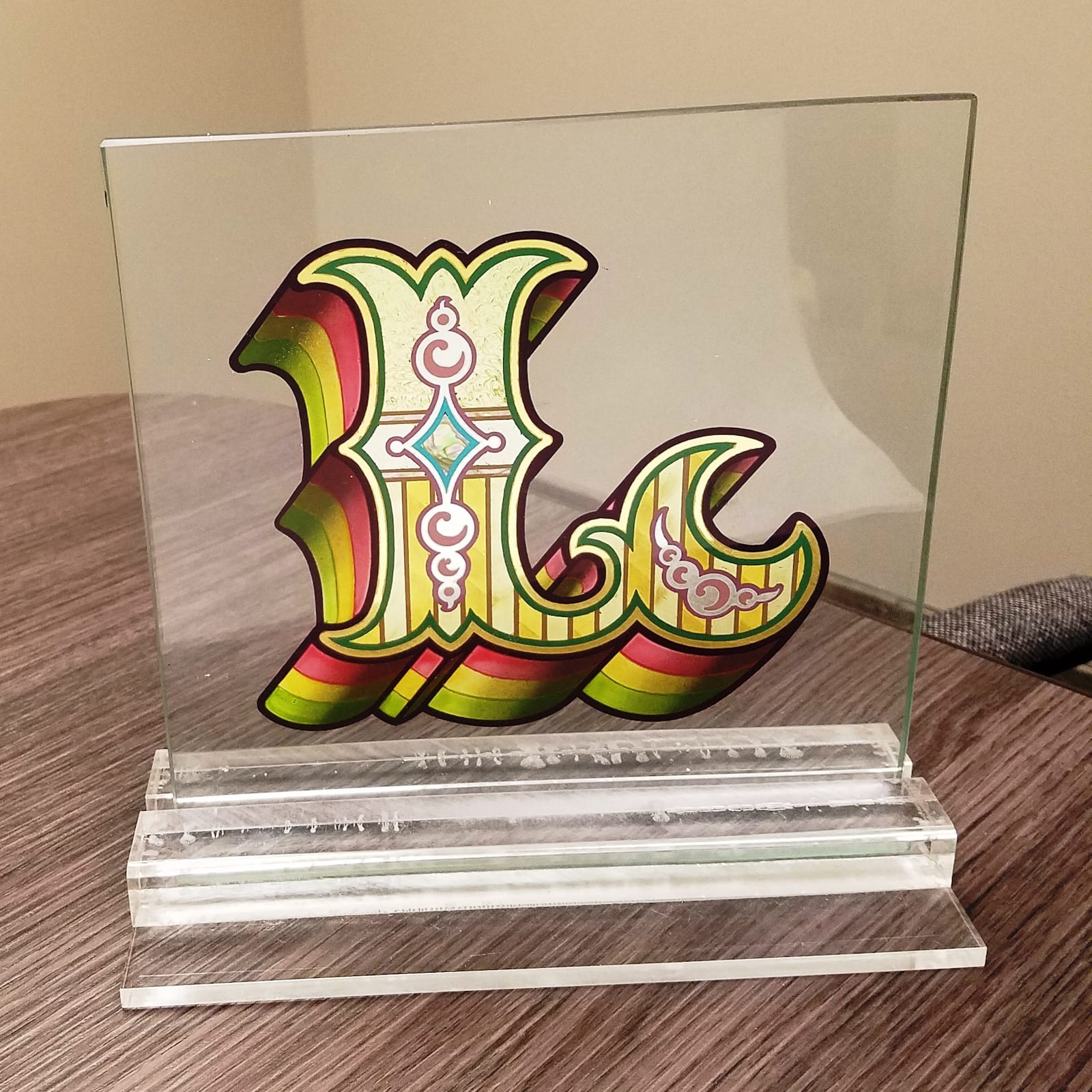 Square panel of glass mounted on a perspex base. The glass is decorated with a Tuscan letter L in the centre, embellished with a host of gilding and lettering effects.
