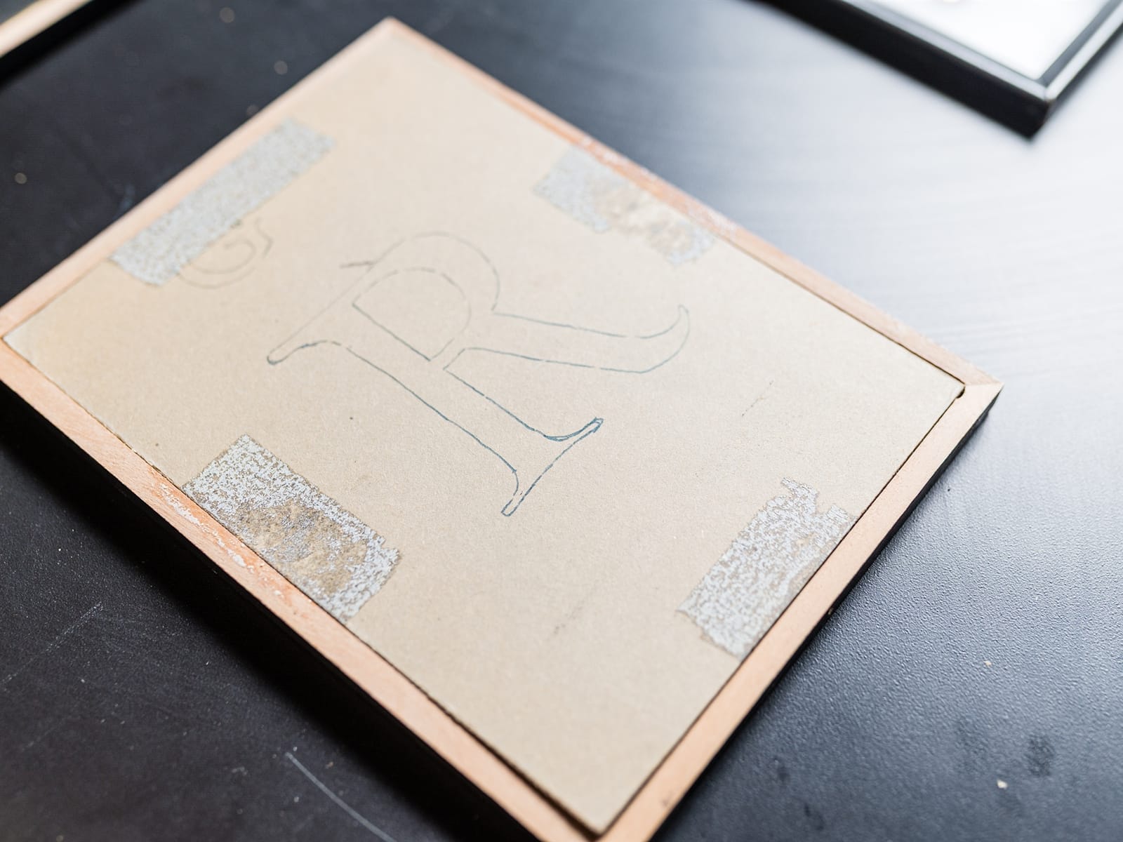 The back side of one of the framed letters with a piece of card set in the portrait oriented frame. A Roman R is drawn in pencil in the middle of the light grey card.