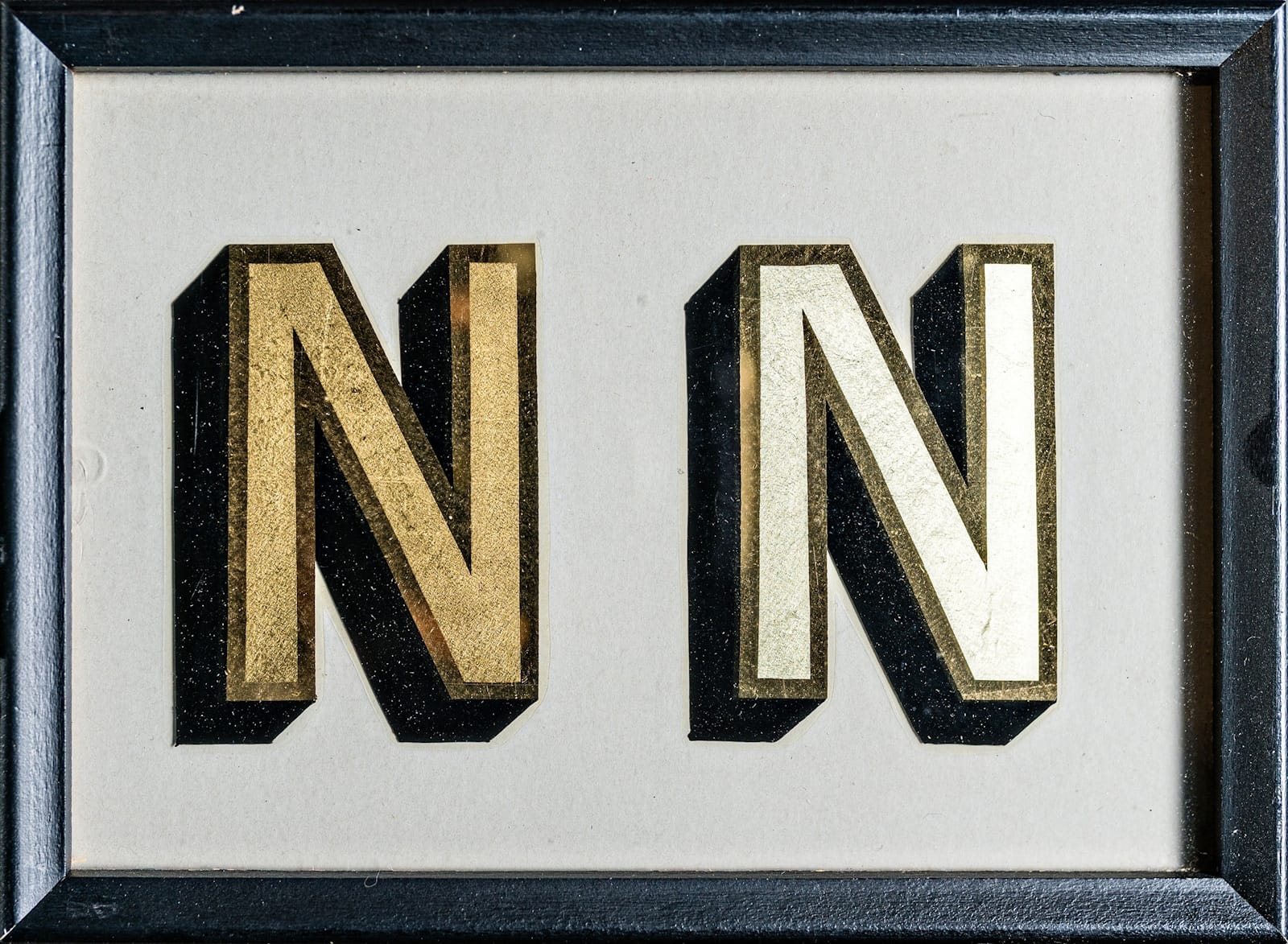 Two san-serif N's, each with a matte gold leaf effect in the centre, outlined with a shiny gild and given a black block shade to the left and down. The one on the left has a darker tone of gold to the one on the right which is almost white in appearance.