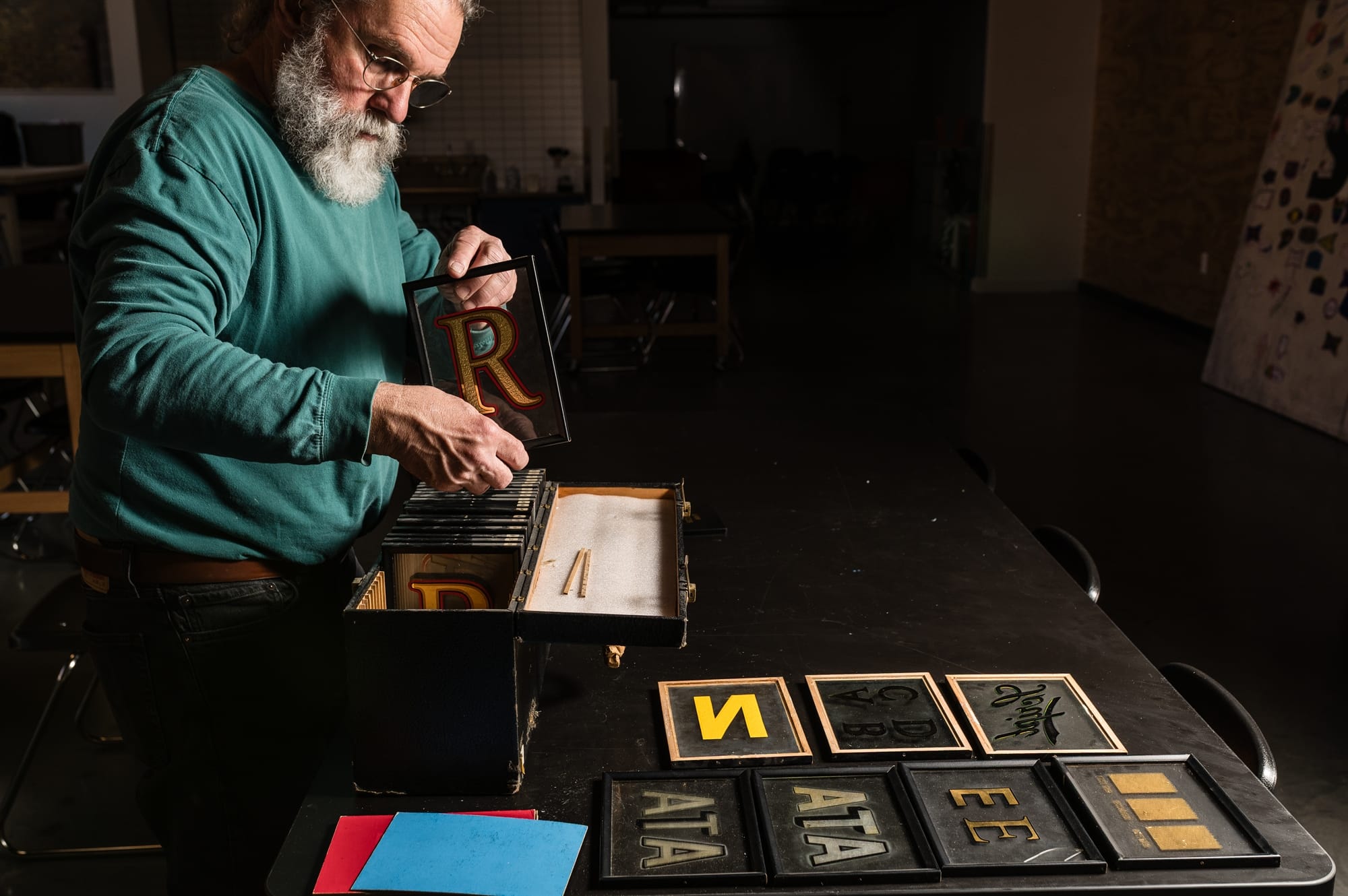 Man holding a framed gilded letter on glass as he removes it from the case to be placed on the dark table alongside others that are already set in an array.