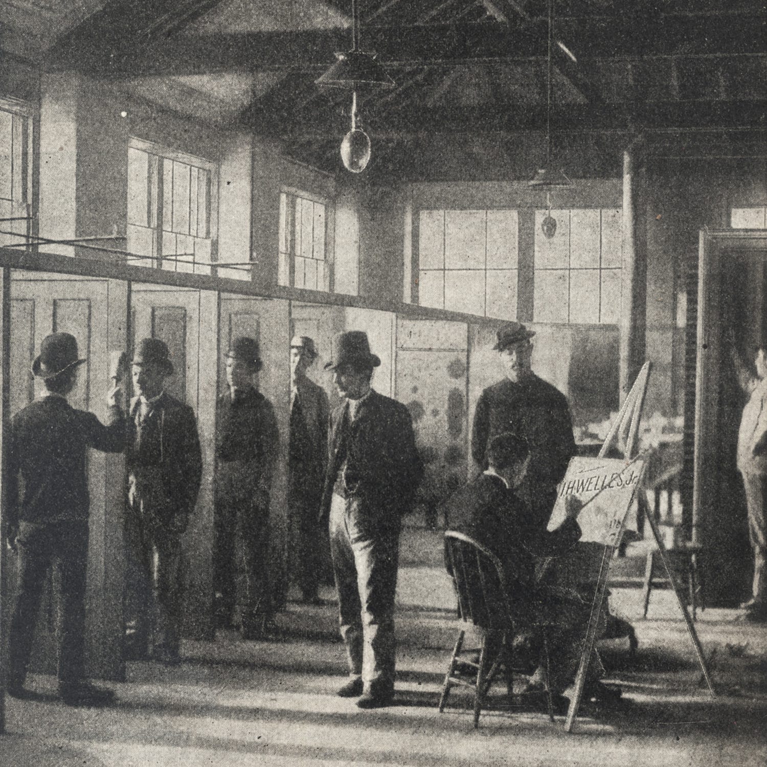 View of a room with a high ceiling and tall windows lettering in lots of light while well-dressed men, predominantly standing, work on painting and decorating tasks. One, in the foreground, is sat at an easel lettering a panel, while others wander around inspecting the work.
