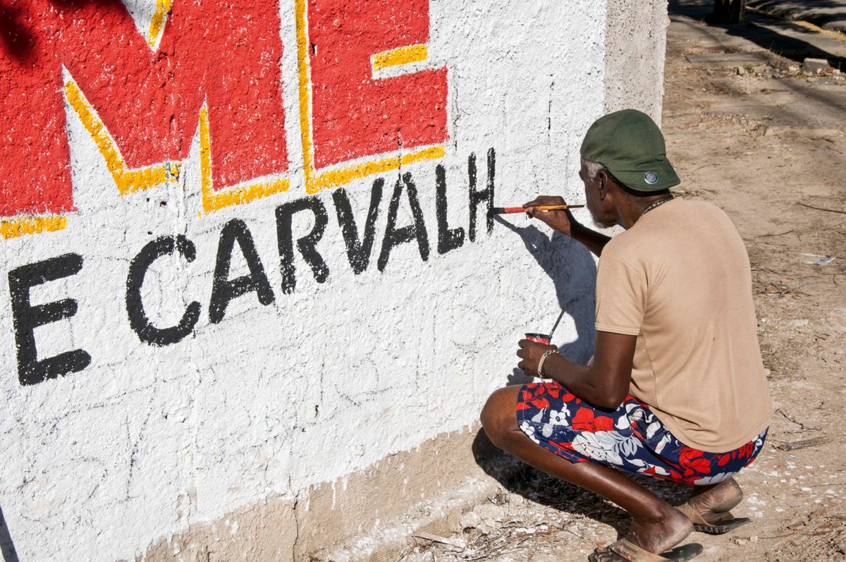 Man crouching in front of a white wall, painting black lettering onto a mural sign.