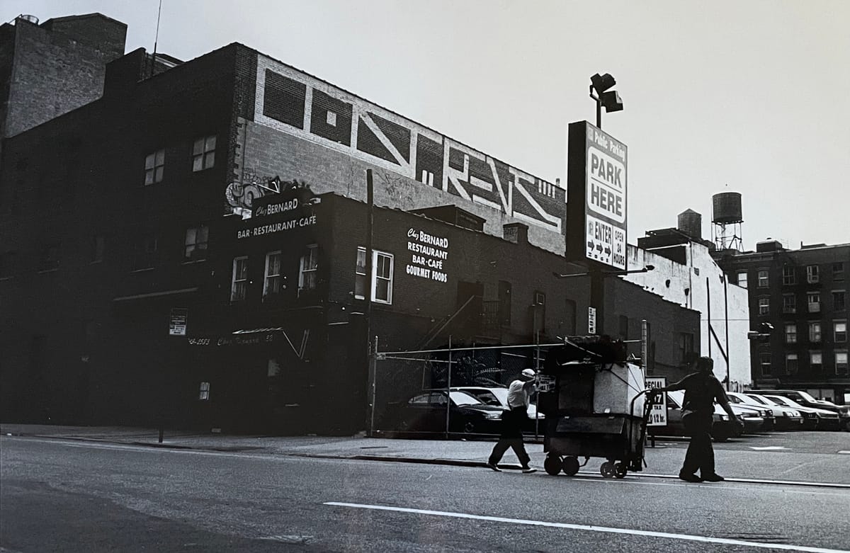Black and white photo of New York street with signs and graffiti on buildings behind a parking lot and men moving a cart.