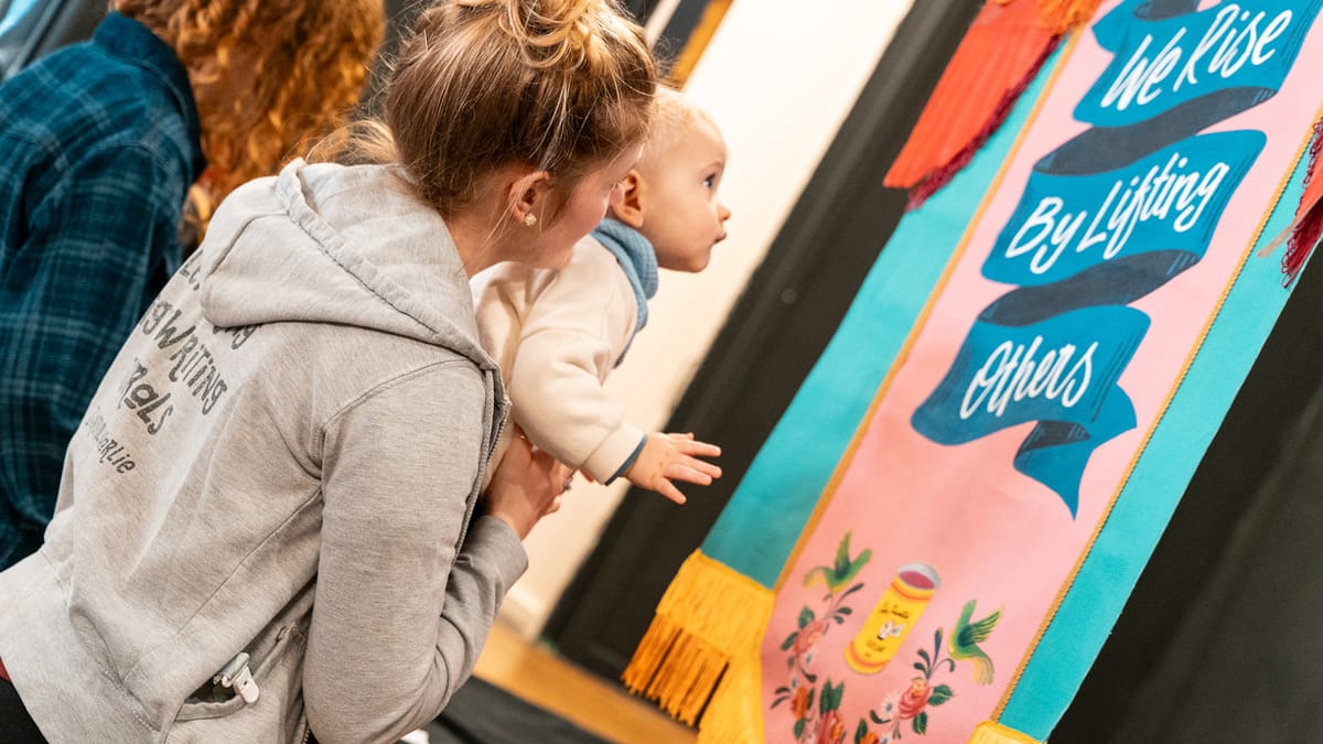 A woman holding a baby that's looking at painted lettering that says 'we rise by lifting others'.