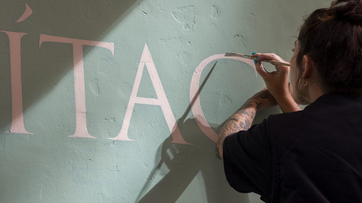 Woman painting pink Roman letters onto a light olive-green wall.