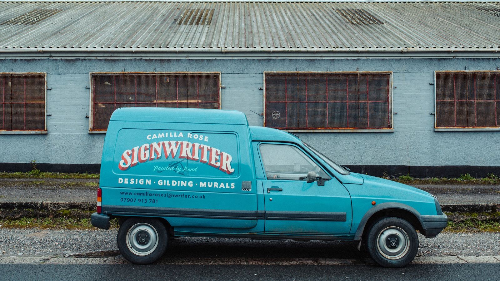 Turquoise van with hand-painted lettering on the side, parked in front of a warehouse-style building.