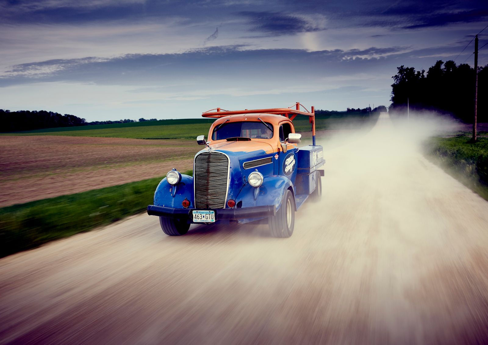 Vintage car painted orange and blue driving down a country road.