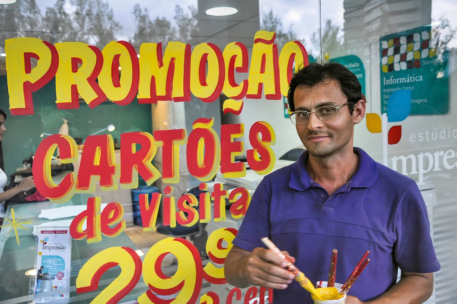 Man posing with paint pot and paint brushes in front of lettering being painted in yellow and red on a window behind him.