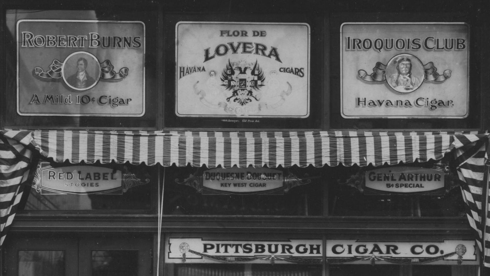 Detail of painted and gilded glass panels advertising cigar brands above the entrance and window of a shop. 