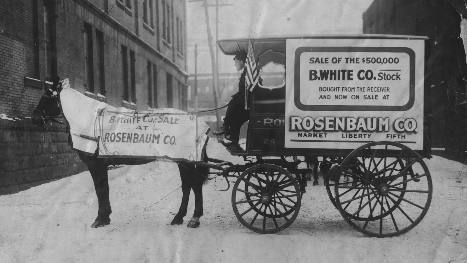 Horse-drawn wagon with driver in the snow. The horse and the wagon are adorned with signage advertising a stock clearance sale.