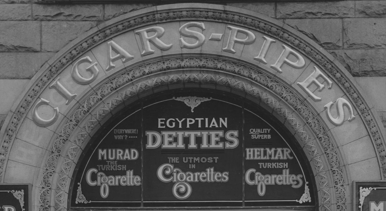 Letters saying "Cigars-Pipes" mounted on an architectural arch, and then signage advertising cigarettes set within the central semi-circle.