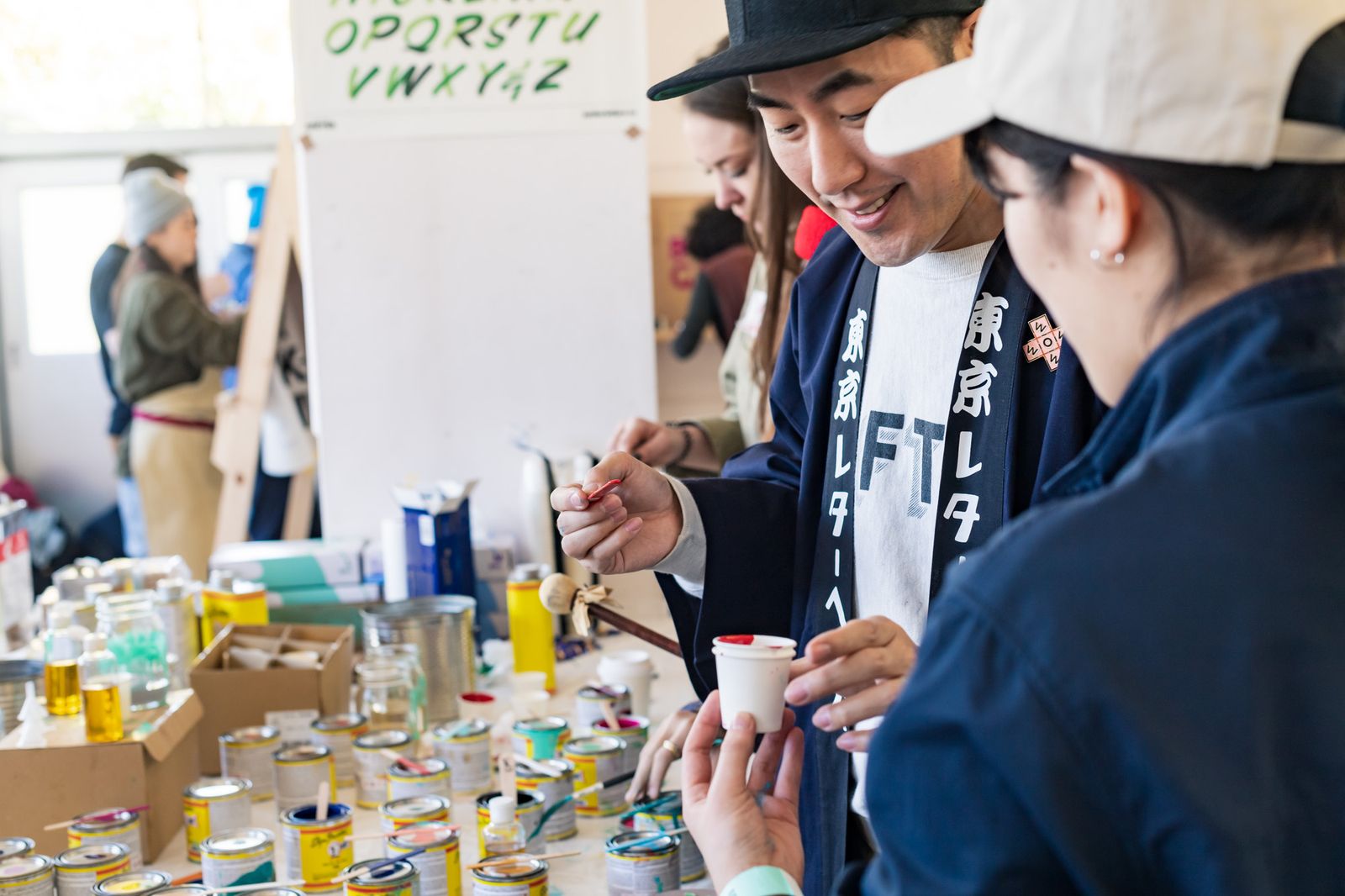 Two people looking at a paper cup with red paint in it, with a table of sign painting paint tins in the background.