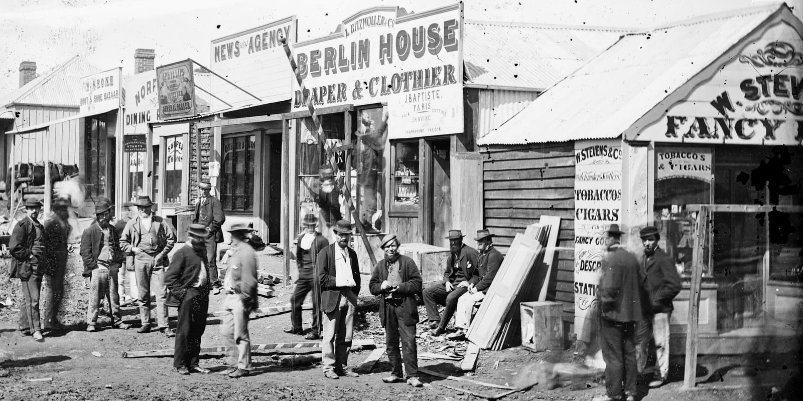 Row of wooden structures housing commercial premises and adorned with hand-painted signs. A number of men are posing of a photo on the rough dirt road in front.