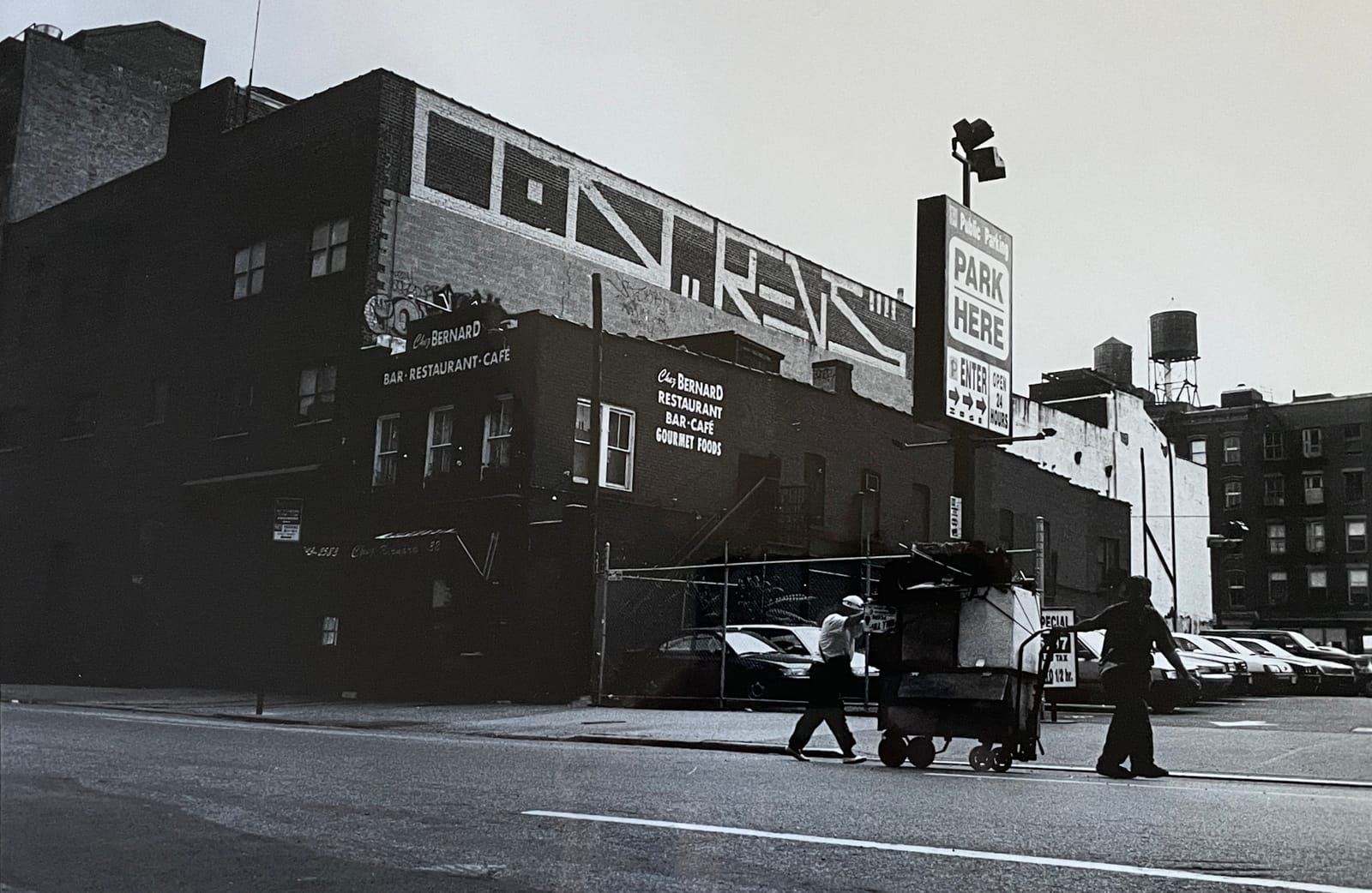 Black and white photo showing a New York street scene with signage and graffiti visible on buildings behind a parking lot and men moving a cart along a road.