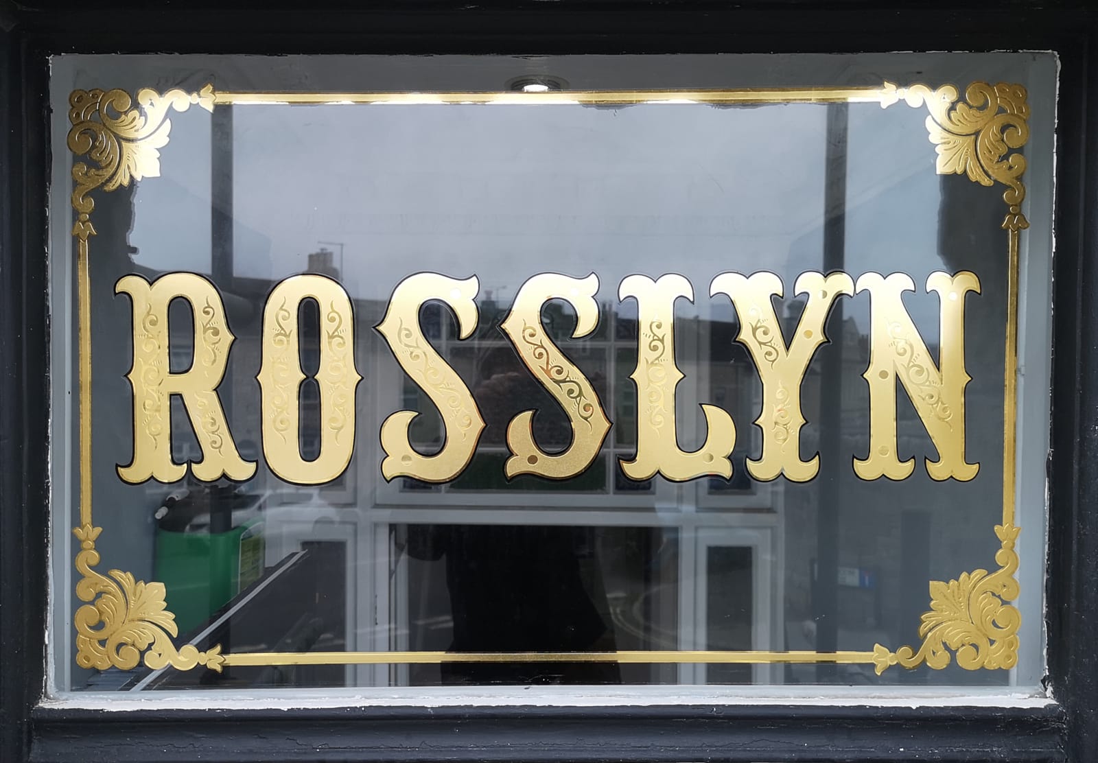 A glass window with gold leaf applied to form an decorative border with corner ornaments and bifurcated lettering in the middle that reads "Rosslyn".