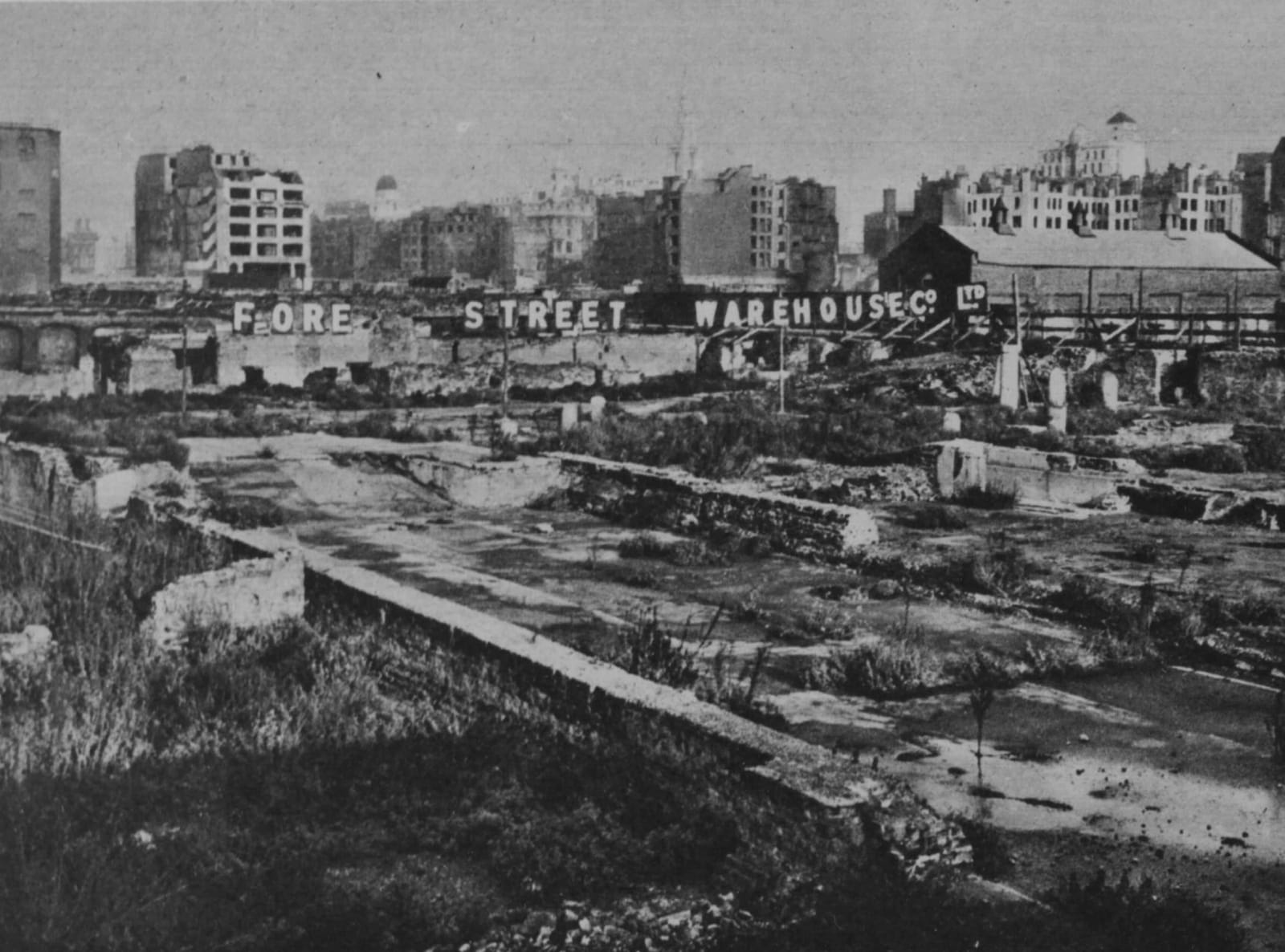 Large expanse of wasteland with bbuildings in the far distance. Large letters are mounted horizontally across a portion of the wasteland, reading "Fore Street Warehouse Co. Ltd."