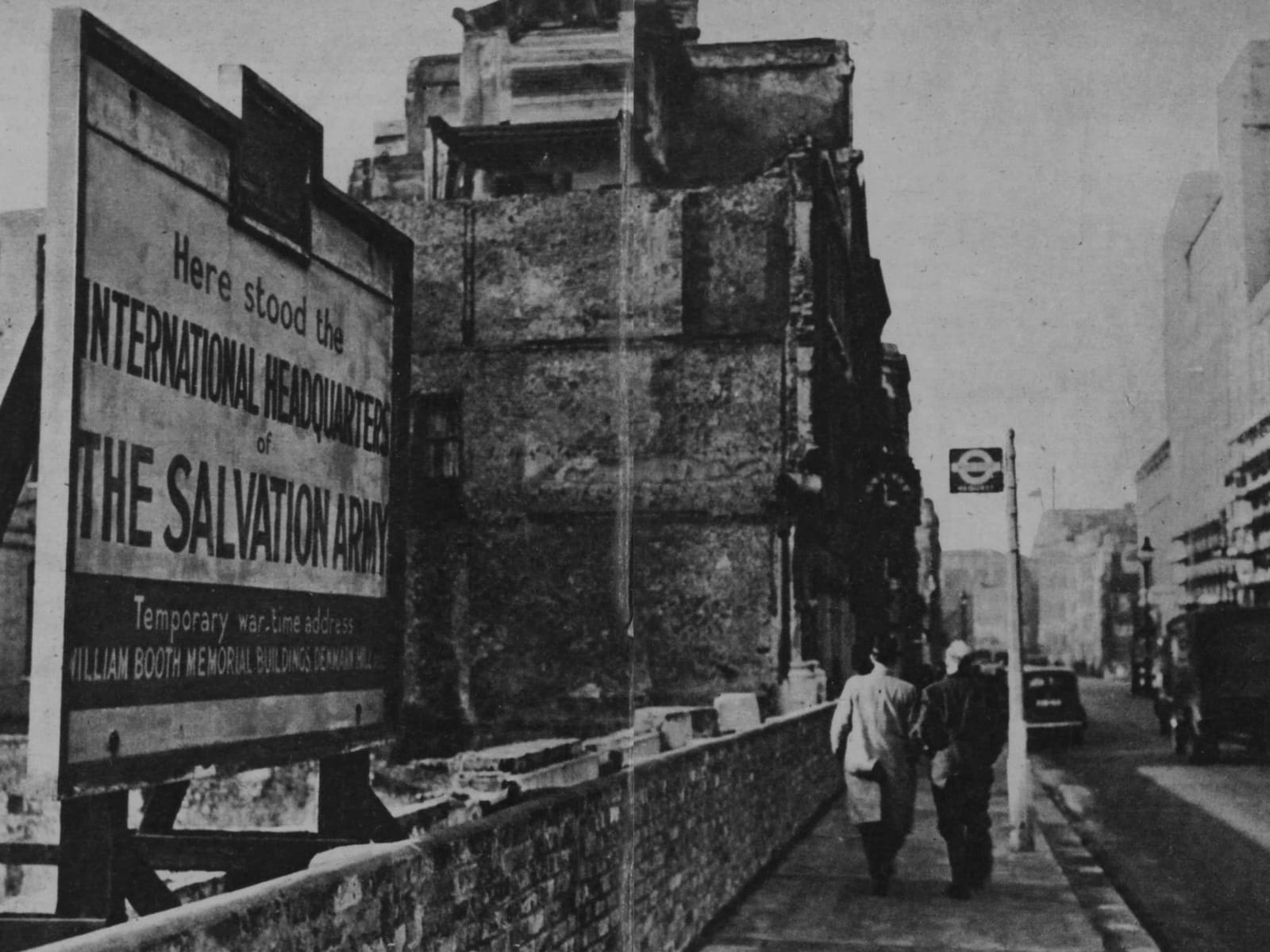 View down a London Street with most buildings still standing. To the left is an empty plot of land with a sign mounted in front reading, "Here stood the international headquarters of The Salvation Army. Temporary war-time address: William Booth Memorial Buildings, Denmark Hill."