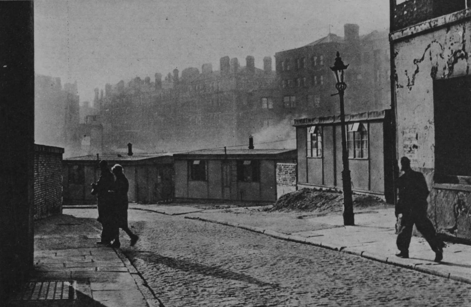 A desolate cobbled street with small bungalow homes sitting in front of larger buildings standing in the distance.