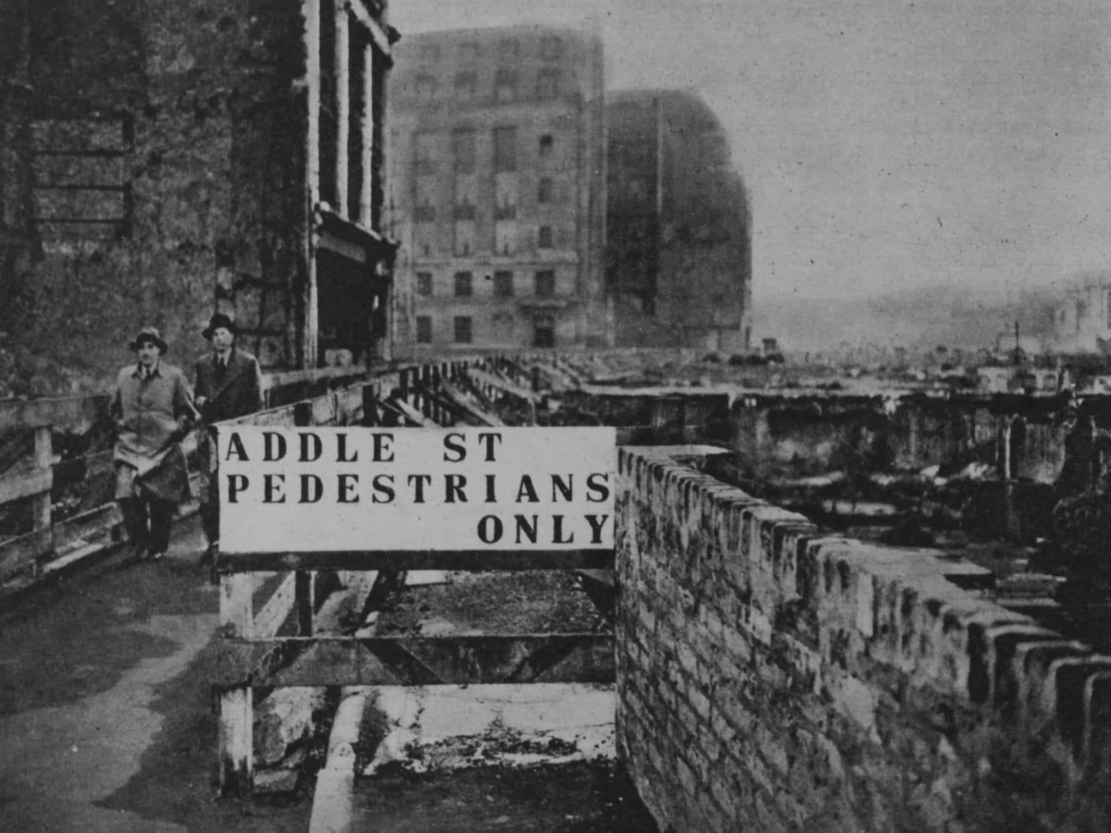 A stencilled sign that reads "Addle St, Pedestrians Only" in front of destroyed and damaged buildings to the right and ahead. There are two men coming towards the camera on the walkway adjacent to the sign.
