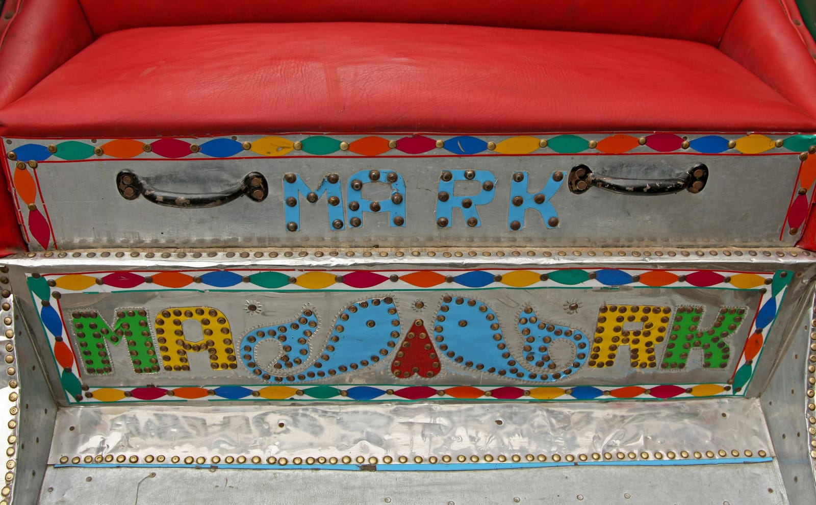Cushioned red seat in a vehicle for transporting passengers. Below this, using a combination of rivets and paintwork, are letters that read "Mark" and decorative elements that include two blue fish either side of an upsidedown heart shape.