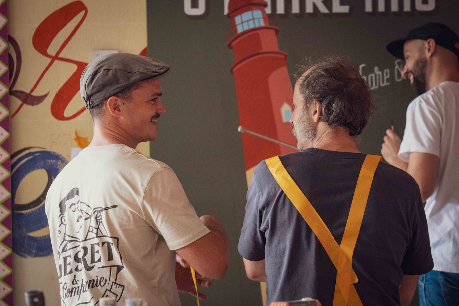 View from behind of two men looking at each other as they talk. In the background another man, slightly out of focus, is pausing from painting a sign, looking to his left and smiling.