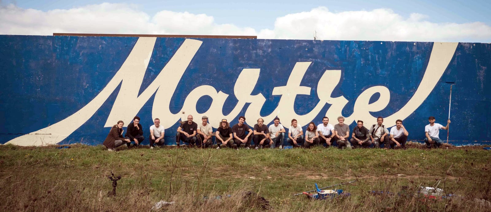 Group of people squatting in front of a large wall that has the word 'Martre' in script lettering produced by cutting in with a dark blue on a white wall.