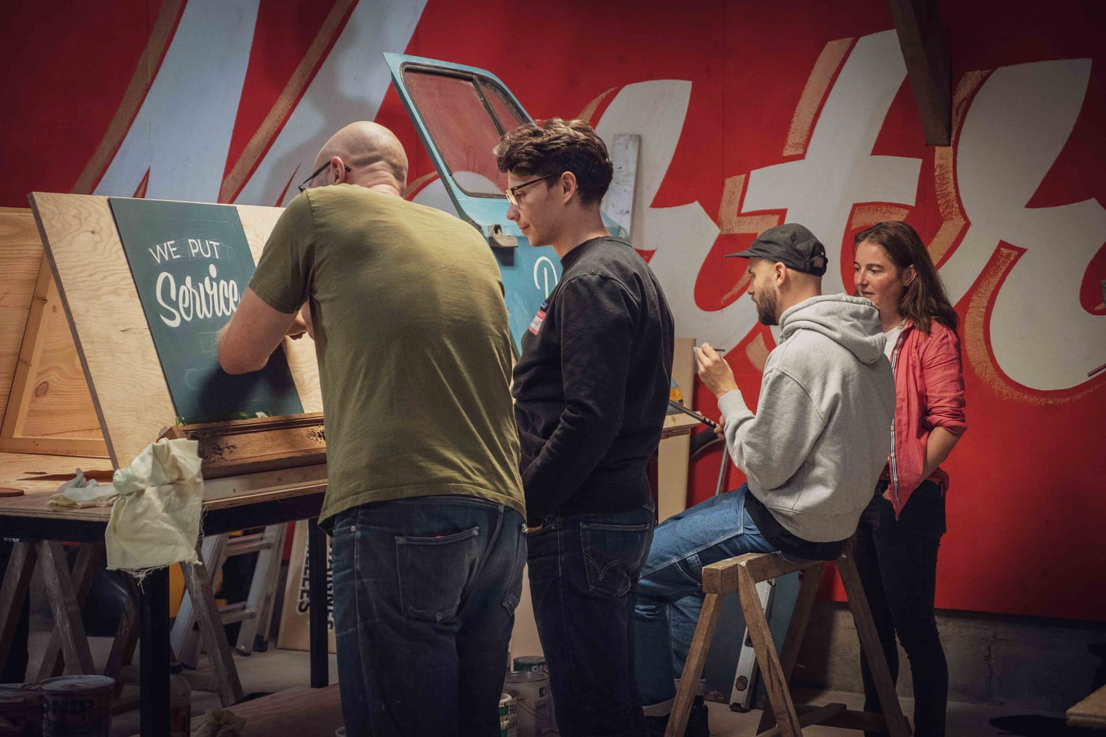 Four people at different points along a wide wooden easel. They are painting letters and talking with each other in pairs.