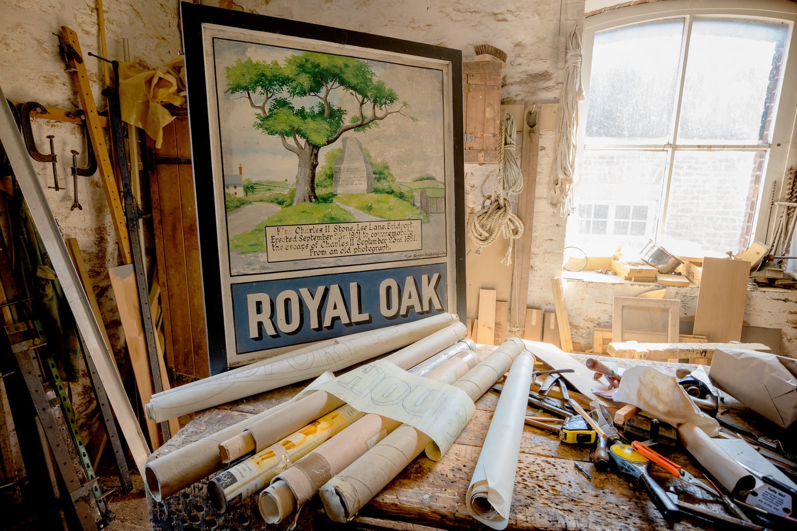 A painted sign for the Royal Oak pub with the name in block letters along the bottom and a pictorial of an oak tree and a rural scene above that. The sign is propped up behind a work bench that is covered with rolls of paper and a variety of tools. Sunlight is streaming in through a window in the background.