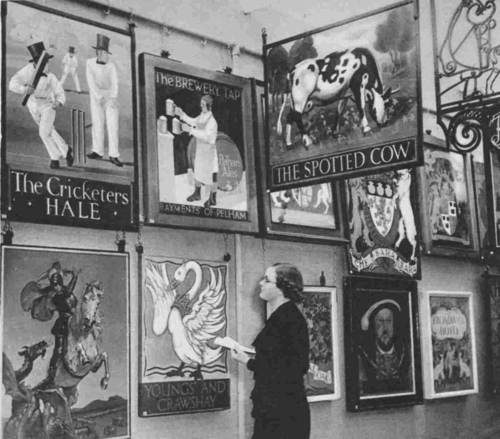 Woman holding an open book and looking at inn signs that are displayed very close to one another in two rows on the wall, while others hang from the ceiling.