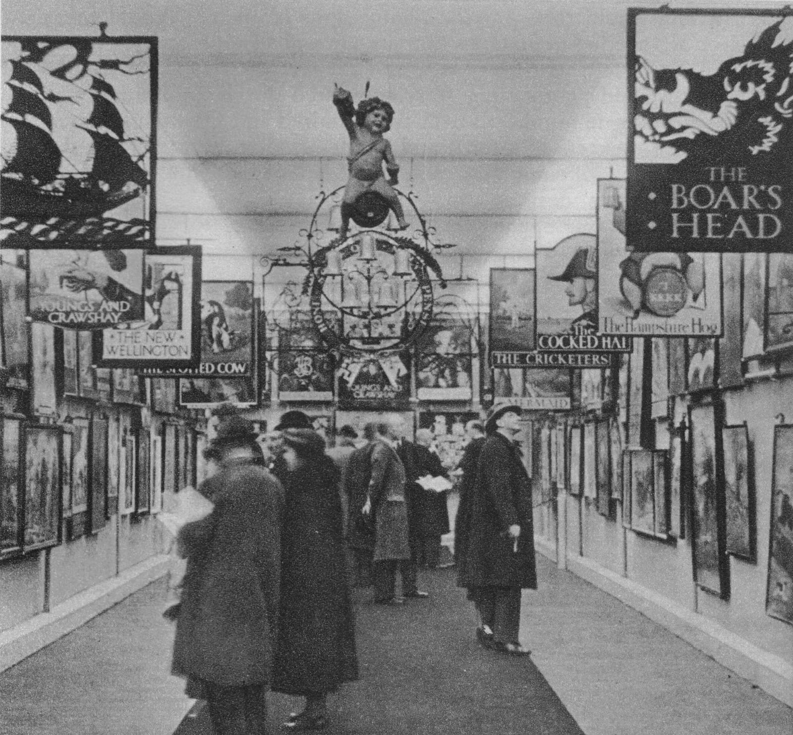 Black and white photo of a gallery setting with smartly dressed people looking at a dense display of inn signs which are mounted to the walls and hanging from the ceiling.